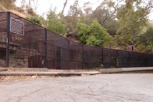 A fenced area with trees and rocky terrain, featuring a sign on the left side and a paved road in the foreground.