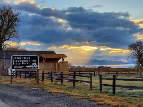 Sign for Camp Floyd Stagecoach Inn State Park at sunrise, with mountains and clouds in the background.