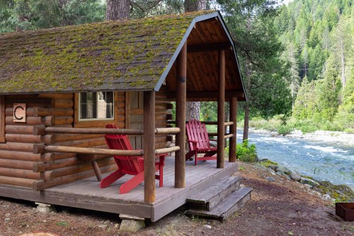 A cozy log cabin with a mossy roof, featuring red chairs on the porch by a flowing river and surrounded by trees.