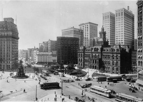 Historic black-and-white photo of a busy city square with buildings, streetcars, and pedestrians.
