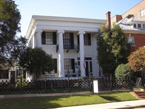 A large white house with columns, black shutters, and a porch, surrounded by greenery and a decorative fence.
