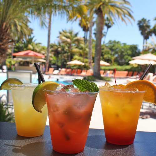 Three colorful cocktails sit on a table, with a pool and palm trees in the background on a sunny day.