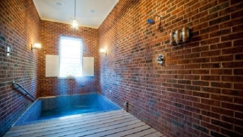 A spacious indoor pool area with brick walls, a window, and a wooden floor, illuminated by wall sconces.