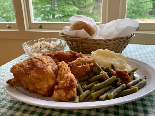 A plate of fried chicken with green beans, mashed potatoes, coleslaw, and a roll in a basket, set on a checkered tablecloth.