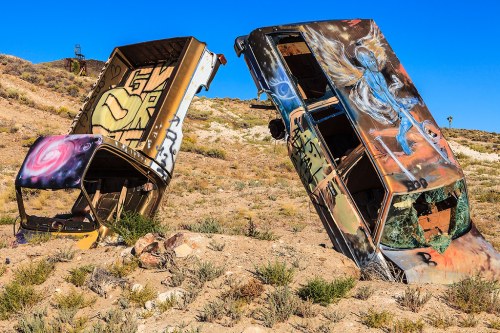 Two abandoned, graffiti-covered vehicles leaning on a hillside, surrounded by dry grass and a clear blue sky.