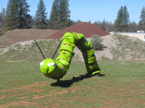 A large green caterpillar sculpture with big eyes, set on a grassy field with trees and a dirt mound in the background.