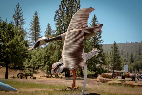 A large metal sculpture of a bird in flight, surrounded by trees and a grassy area.