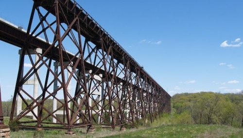 Rusty railway trestle bridge spans a green landscape under a clear blue sky with scattered clouds.