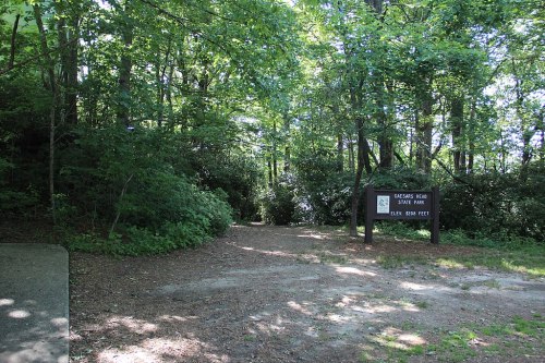 A wooded area with a dirt path leading to a sign for "Oglebay Park" surrounded by lush greenery.