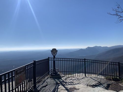 A scenic overlook with a viewing platform, featuring distant mountains under a clear blue sky. A warning sign is visible.