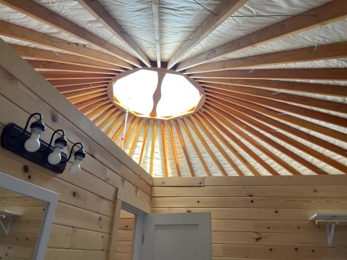 Interior view of a yurt with a circular skylight and wooden beams, featuring a light fixture and wooden walls.