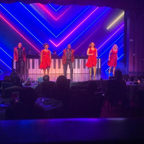 A lively performance on stage with five singers in red dresses, accompanied by musicians at a piano, colorful lights in the background.