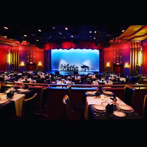 Elegant dining setup in a theater with tables, plates, and a stage featuring a piano and band instruments in the background.