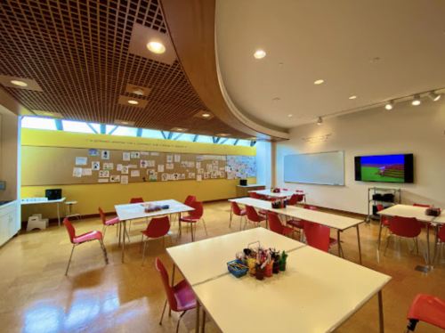 A bright classroom with red chairs, white tables, and a wall covered in student artwork and a TV screen.