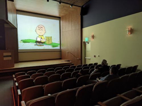 A small theater with two people watching a cartoon featuring a character sitting on a green bench.