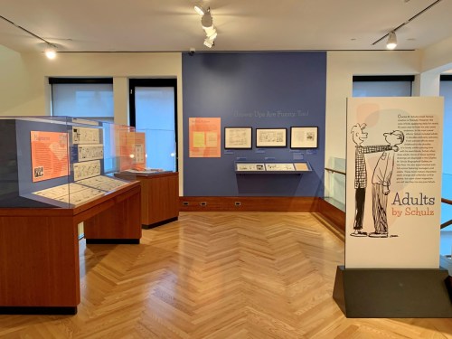 Exhibit featuring comic art and displays about "Adults by Schulz" in a gallery setting.
