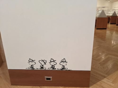Three cartoon characters in hats walking along the base of a white wall in a museum setting.