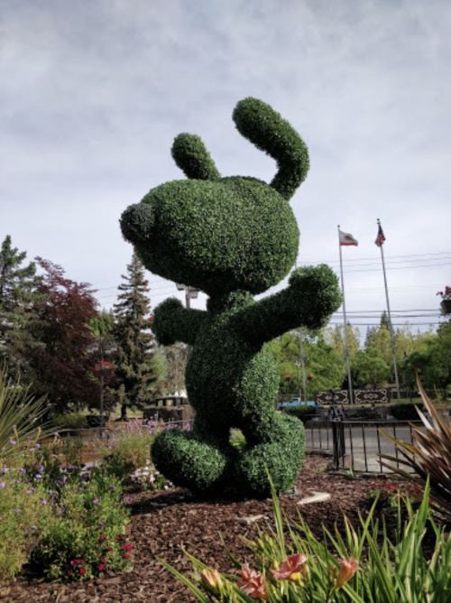 A large topiary sculpture of a cartoon dog with ears, standing in a garden surrounded by flowers and flags.