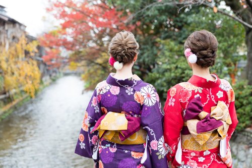 Two women in colorful kimonos stand by a serene river, surrounded by autumn foliage.