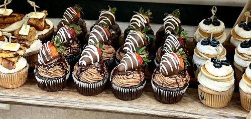 A display of cupcakes topped with chocolate-dipped strawberries and drizzled chocolate, arranged on a wooden shelf.