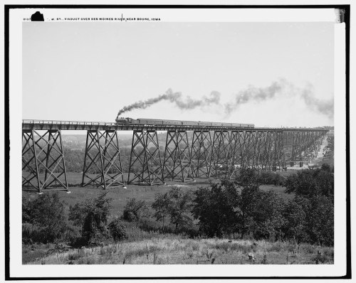 A steam train crosses a tall wooden trestle bridge over the Des Moines River in Iowa, surrounded by greenery.