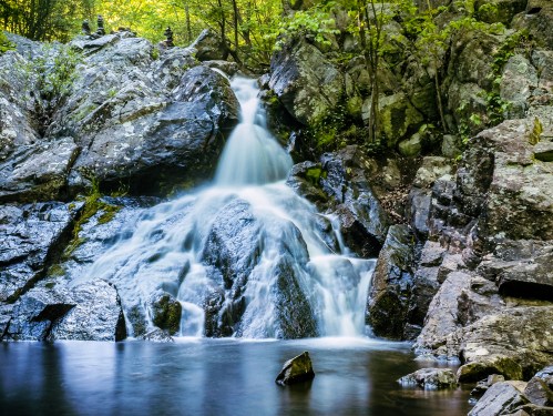 A serene waterfall cascading over rocks into a calm pool, surrounded by lush greenery and natural stone formations.