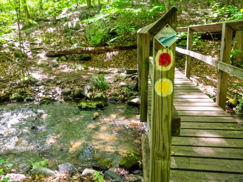 A wooden bridge over a stream in a lush, green forest, marked with red and yellow trail signs.