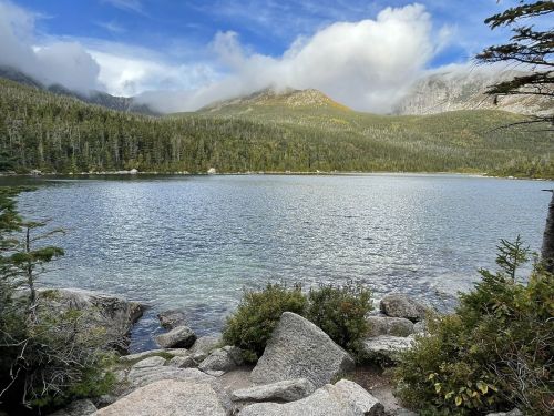 A serene lake surrounded by lush forests and mountains under a partly cloudy sky. Rocks line the shore.