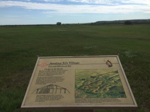 Sign at Awitxa Xí Village site, with grassy landscape and distant hills under a clear blue sky.