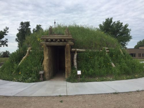A grassy, earthen structure with a wooden entrance, surrounded by trees and a paved walkway.