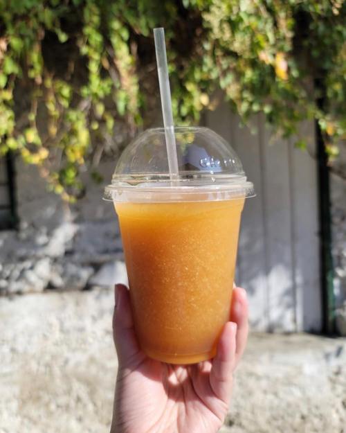 A hand holding a clear cup of orange slush with a straw, set against a backdrop of greenery.