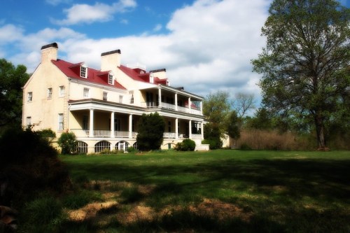 A large, historic house with a red roof, surrounded by green grass and trees under a partly cloudy sky.