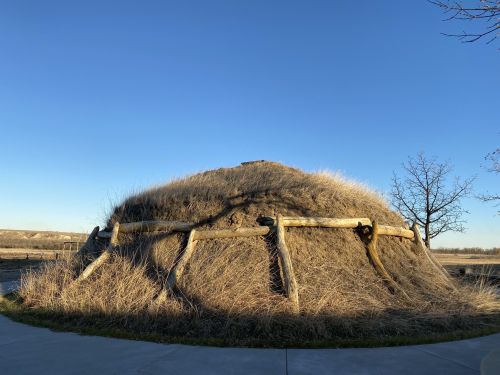 A grassy, dome-shaped structure with wooden supports, set against a clear blue sky and sparse trees.