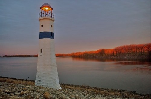 A lighthouse stands by a calm river at dusk, with trees reflecting warm colors in the background.