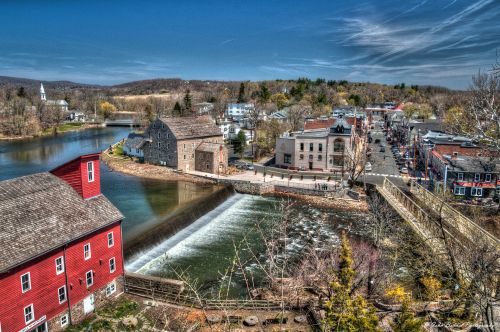 A scenic view of a small town with a river, a waterfall, and historic buildings under a clear blue sky.