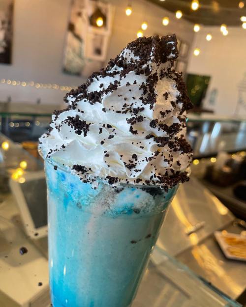 A tall cup of blue milkshake topped with whipped cream and crushed cookies, set in a cozy café environment.