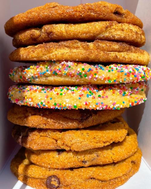A stack of colorful cookies, including chocolate chip and sprinkle-covered varieties, in a white box.