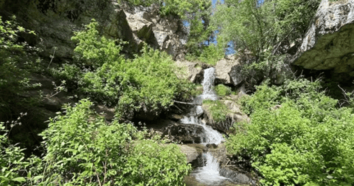 A serene waterfall cascading down rocky terrain, surrounded by lush green foliage and trees under a clear blue sky.