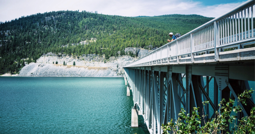 A long bridge extends over a turquoise lake, surrounded by green hills and rocky cliffs under a clear sky.