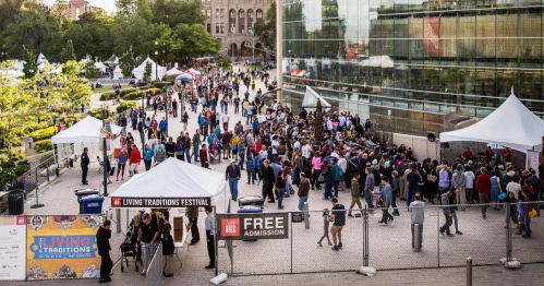 A large crowd gathers at the Living Traditions Festival, with tents and a modern building in the background.