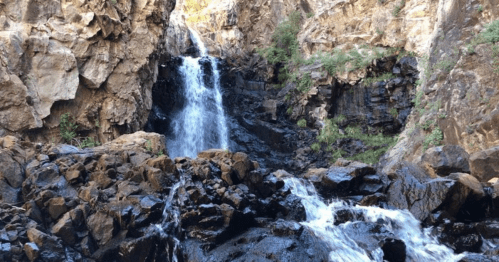 A cascading waterfall flows over rocky cliffs, surrounded by lush greenery and rugged terrain.