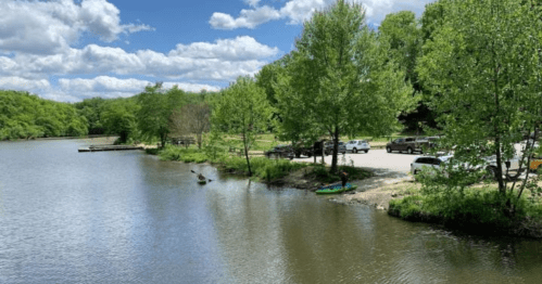 A serene river scene with green trees, a parking area, and a kayak near the shore under a partly cloudy sky.