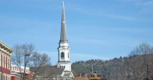 A tall church steeple rises against a clear blue sky, surrounded by trees and distant mountains.