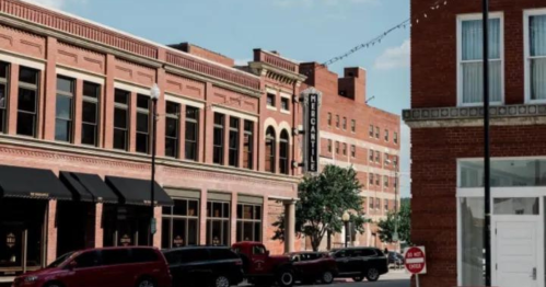 Historic brick buildings line a street, with parked cars and trees, under a clear blue sky.