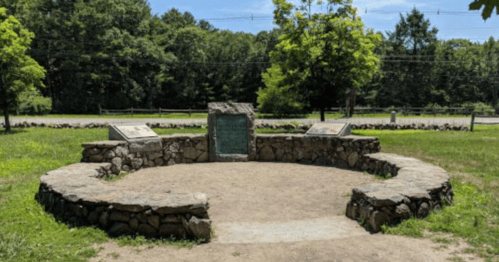 A circular stone monument in a grassy area, surrounded by trees, with informational plaques on either side.