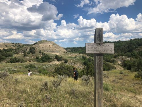 A wooden sign points to nearby caves, with a hilly landscape and cloudy sky in the background. Two people walk in the distance.