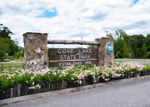 Sign for Cove Lake State Park, established in 1940, surrounded by flowers and greenery under a cloudy sky.
