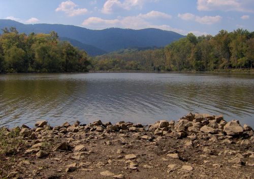 A serene lake surrounded by trees and mountains under a partly cloudy sky, with rocky shore in the foreground.