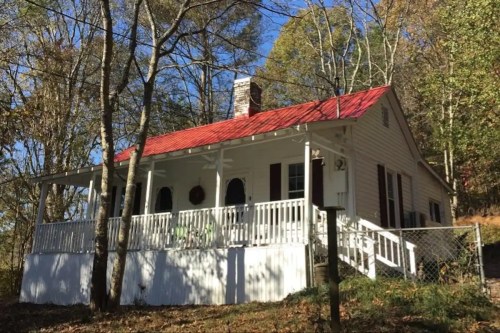 A charming white house with a red roof, surrounded by trees and a wooden porch, set in a serene outdoor landscape.