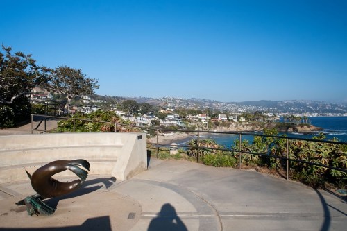 A coastal view featuring a sculpture, greenery, and houses on a hillside under a clear blue sky.